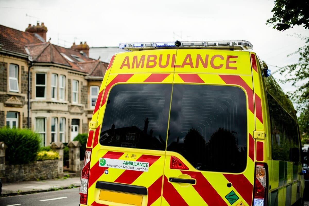 A yellow and red striped ambulance with emergency medical services stickers is parked on a residential street lined with stone houses and greenery under a cloudy sky.